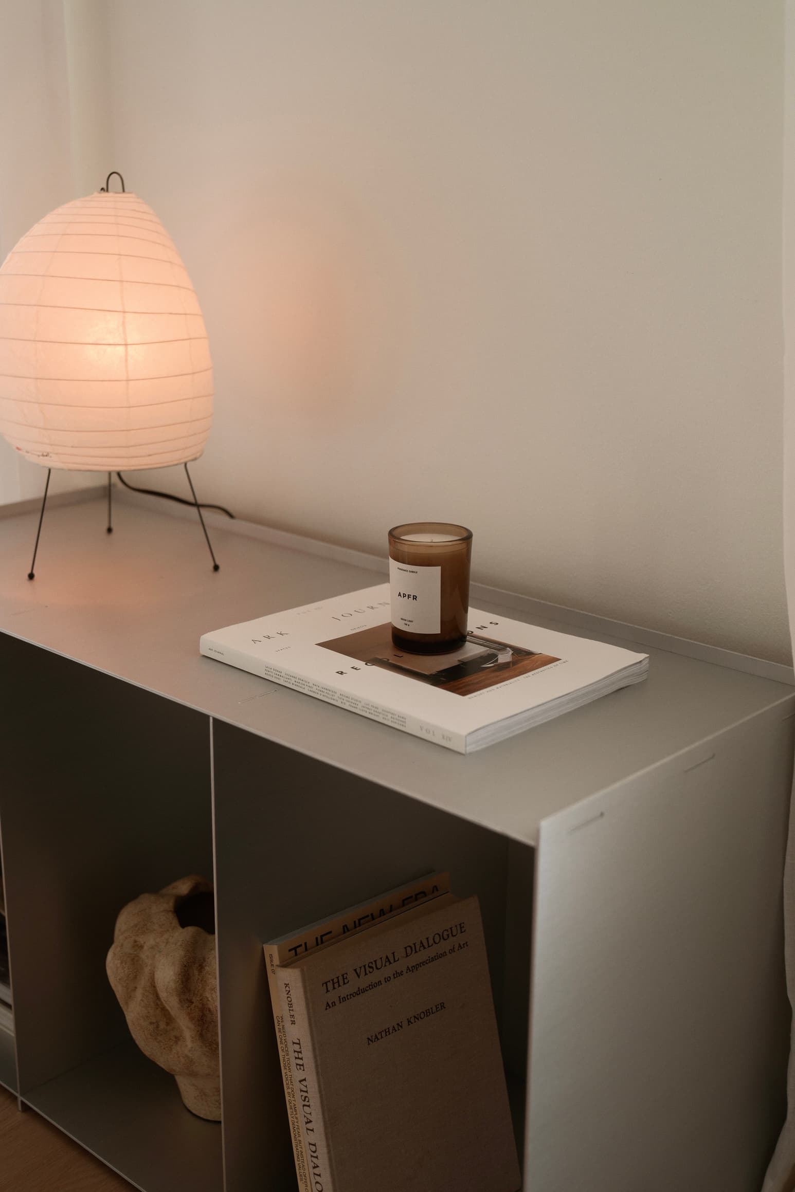 A paper lantern lamp glowing on a white sideboard, beside a candle and a magazine